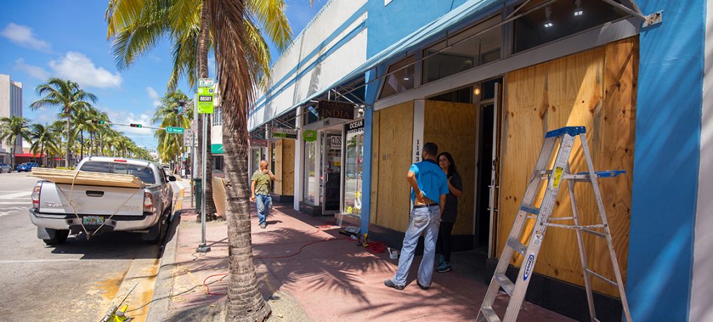 Photo of a couple talking in front of a boarded up business with palm trees and a parked truck on the road.