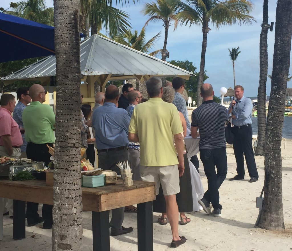 Photo of a man speaking to a group of people outside on a beach.