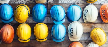 Photo of blue, yellow, orange and white hardhats hanging on a wooden wall.