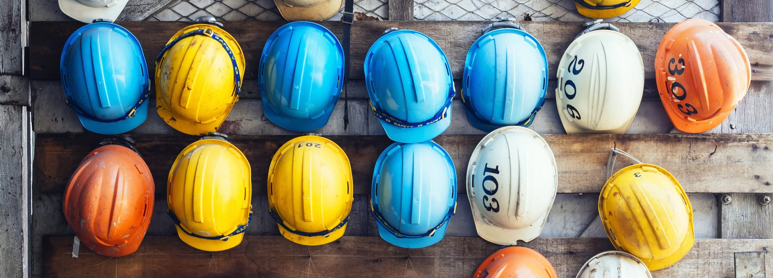 Photo of blue, yellow, orange and white hardhats hanging on a wooden wall.